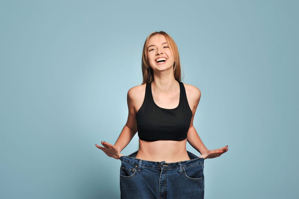 Happy woman showing weight loss success Joyful woman in black sports bra with loose jeans, celebrating weight loss achievement.